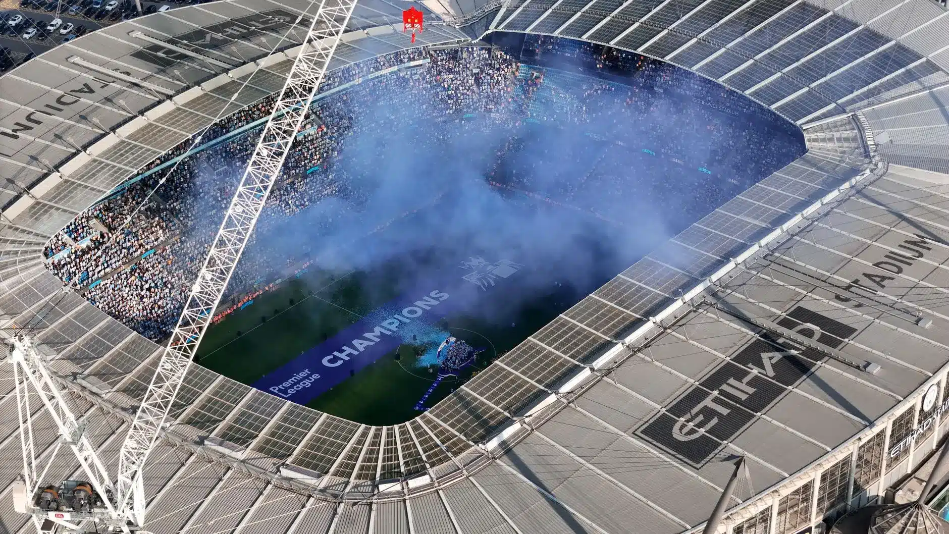 High-angle view of Etihad Stadium with a blue smoke display and a Premier League Champions banner on the pitch.